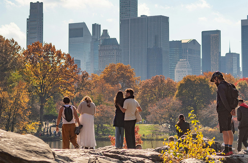 People standing on a rock in Central Park, looking out over lush green trees toward the towering skyscrapers of the Midtown Manhattan skyline under a hazy