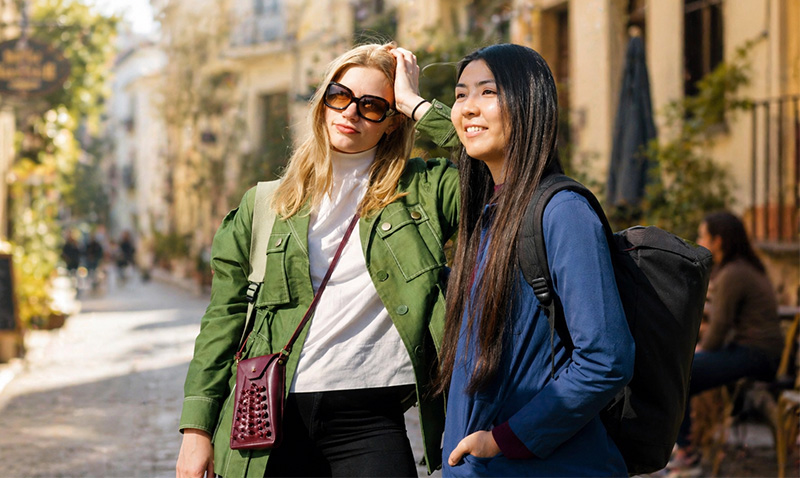 Two women standing together on a cobblestone street in Europe. One woman wears a green puffer vest over a white hoodie and sunglasses, while the other wears a dark blue denim jacket and a crossbody bag. Historical European architecture and a yellow tram are visible in the background.