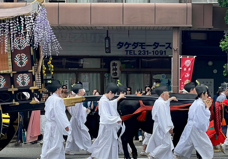 A group of people dressed in traditional white Shinto robes and black hats parading through a Kyoto street during a festival, with a decorated float and traditional Japanese storefronts in the background.