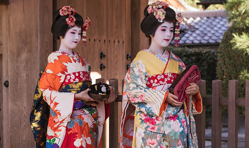 A detailed view of a geiko and maiko standing together in front of a traditional wooden machiya house in Kyoto, showcasing the contrast between the maiko’s colorful kimono and long dangling obi and the geiko’s more understated, elegant black kimono.