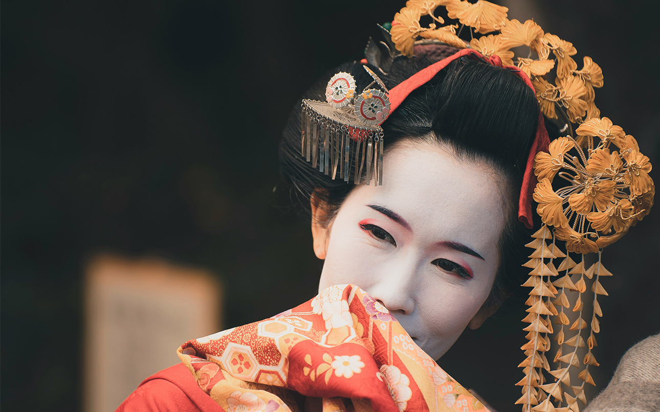 A close-up portrait of a geiko (geisha) in Kyoto, featuring traditional white oshiroi makeup, elaborate hair ornaments (kanzashi), and a vibrant red kimono with intricate floral patterns.