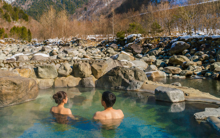 Couple relaxing in a natural outdoor hot spring surrounded by rocky terrain and snow-covered mountains under a clear blue sky in Japan