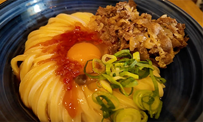 A bowl of kama-tama udon topped with a raw egg yolk, green onions, and tempura bits, representing a comforting first meal in Tokyo.