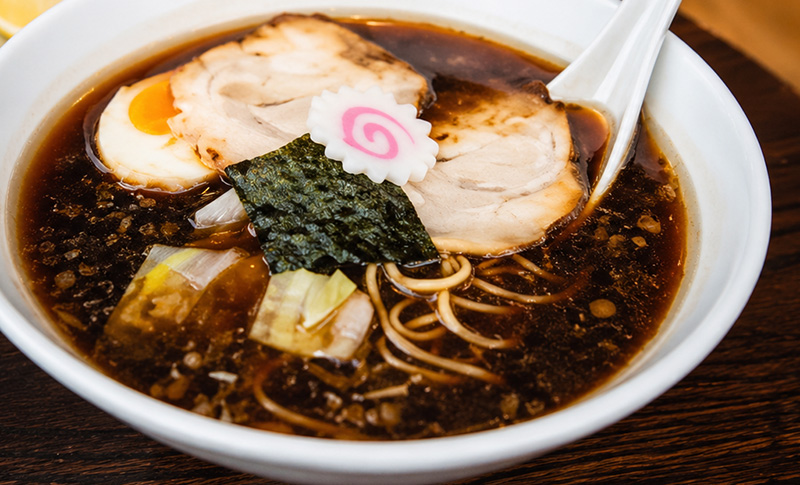 Close-up of a rich Kyoto-style ramen bowl with dark broth, sliced pork, soft egg, seaweed, and noodles in a clean white bowl on a wooden table.