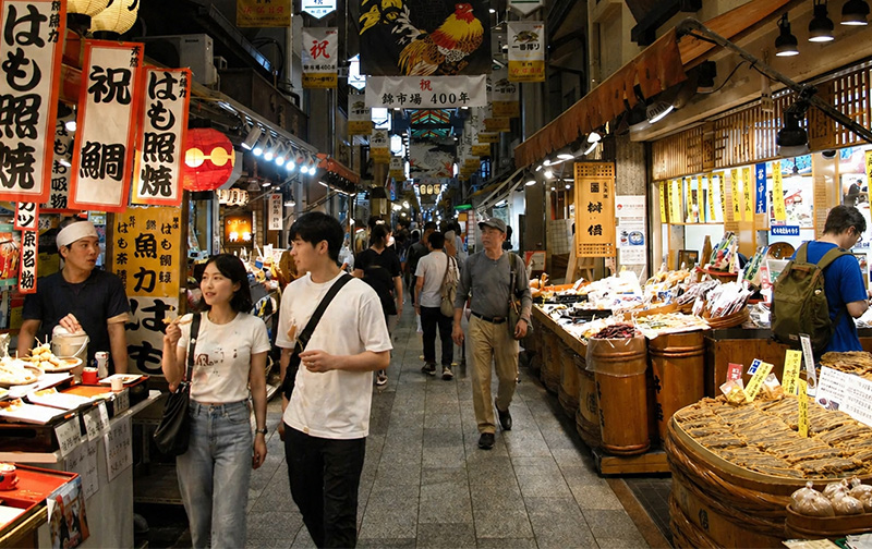 Bustling indoor food market in Kyoto with narrow walking lane, traditional lanterns, colorful overhead panels, and vendors serving local street food as shoppers browse stalls on both sides.