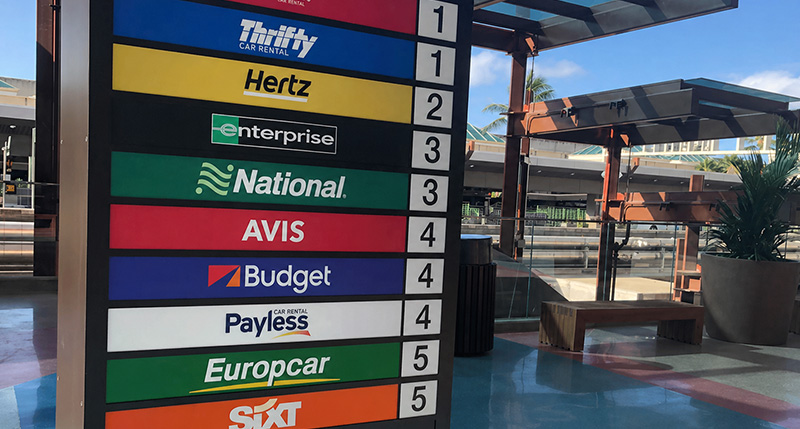 A colorful airport directional sign listing various rental car agencies including Thrifty, Hertz, Enterprise, National, Avis, Budget, Payless, Europcar, and Sixt. Each brand is associated with a specific terminal floor number.