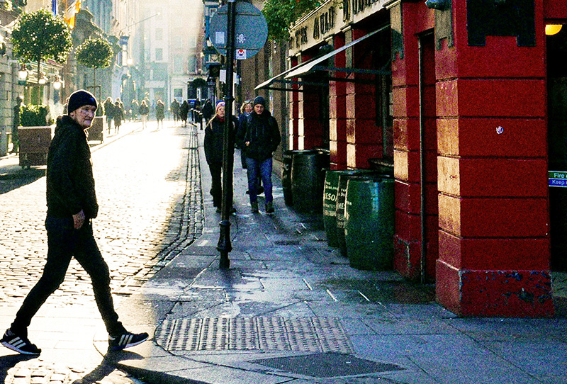 A man in a dark jacket and jeans walks past a row of traditional red-fronted shops and pubs on a narrow, cobblestone street in Dublin’s Temple Bar district.