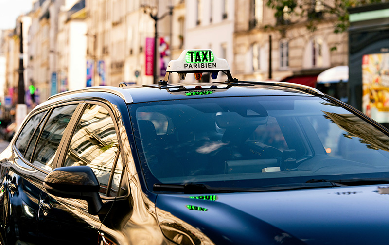 A close-up shot of a Paris taxi's roof sign and side mirror in a busy urban setting, with city traffic blurred in the background.