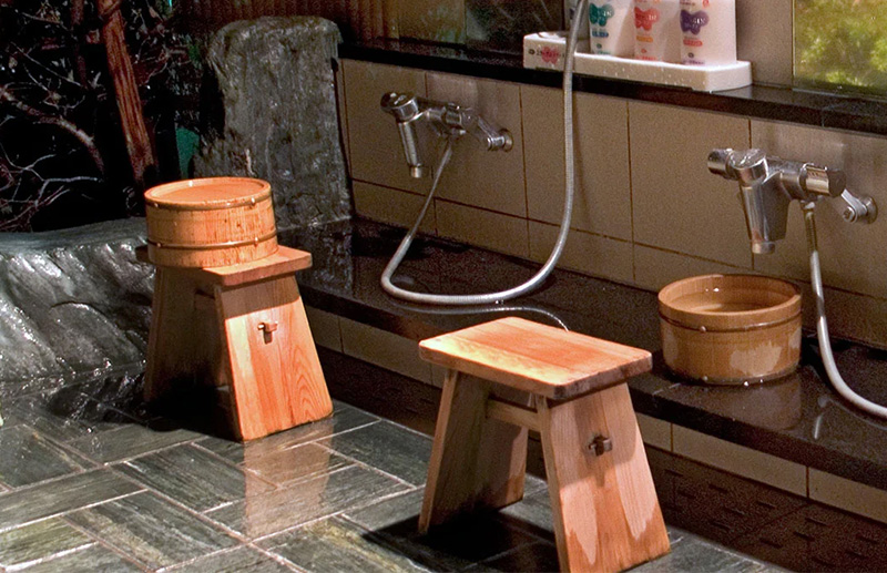A traditional Japanese indoor washing station with wooden buckets, low stools, and hand-held showers used for cleaning before entering an onsen bath.