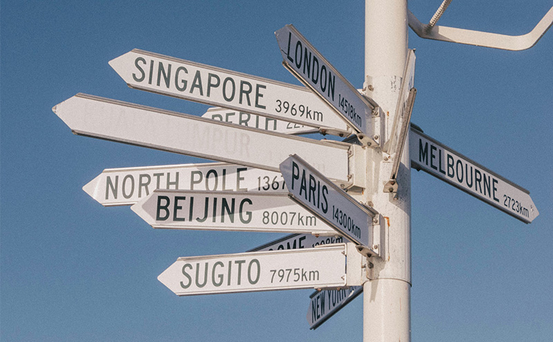 A wooden directional signpost against a blue sky, with arrows pointing to various global cities including Singapore, London, Beijing, Paris, and Tokyo, along with their respective distances in kilometers.