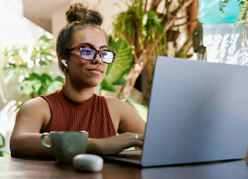 A woman wearing glasses and a sleeveless top sits at a table, focused on her laptop while wearing earbuds in a bright, modern indoor space.