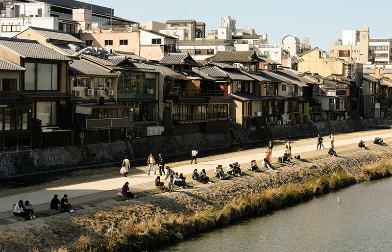 A peaceful landscape of the Kamogawa River in Kyoto at dusk, featuring the iconic wooden dining platforms (yuka) of restaurants overlooking the water with soft evening lighting and the mountains in the distance.