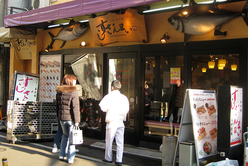 Exterior of a Sushizanmai restaurant in Tokyo featuring two large, realistic tuna sculptures mounted above the entrance and a chef in a white uniform standing outside