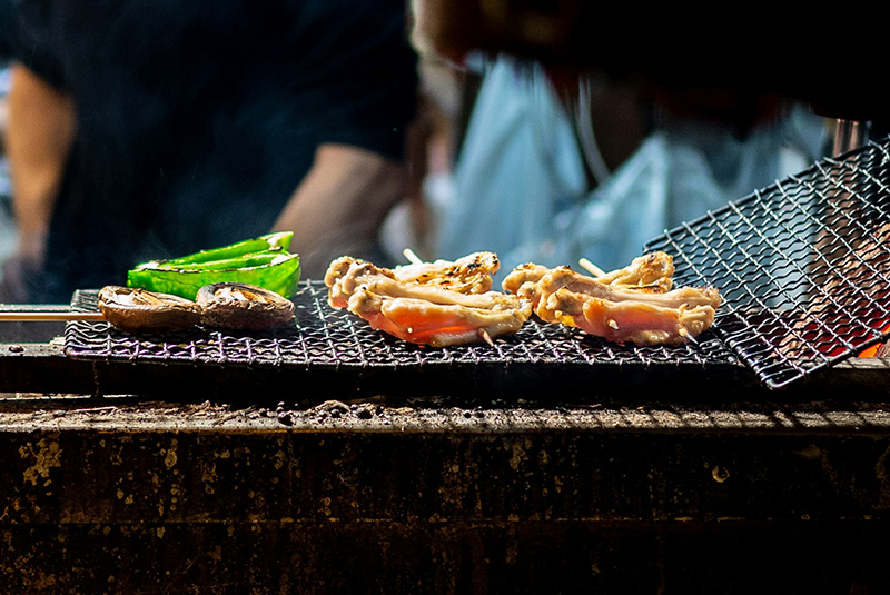 A close-up shot of a chef's hands using metal tongs to grill skewers of yakitori over an open charcoal flame at a traditional Japanese izakaya in Tokyo.