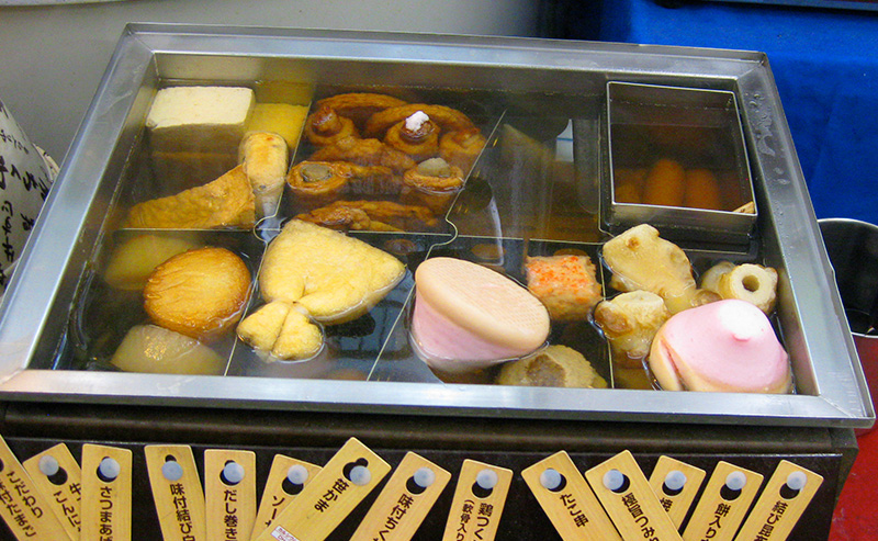 Assorted oden items, including daikon, fish cakes, and tofu, simmering in a partitioned metal broth container at a Tokyo food stall or convenience store.