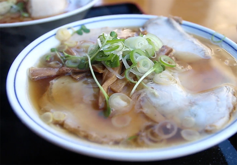 A steaming bowl of Tokyo-style shoyu ramen with chashu pork, bamboo shoots, and green onions, served at a traditional wooden ramen counter.