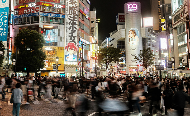 A high-angle view of the Shibuya Scramble Crossing at night, showing hundreds of pedestrians crossing the multi-directional intersection surrounded by towering buildings with bright neon advertisements and large video screens.
