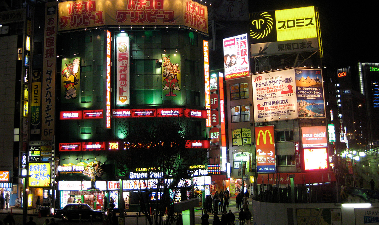 A vibrant night scene in a busy Tokyo district featuring a dense collection of glowing neon restaurant and shop signs in Japanese characters, with a McDonald's logo visible among the illuminated storefronts and pedestrians walking below.