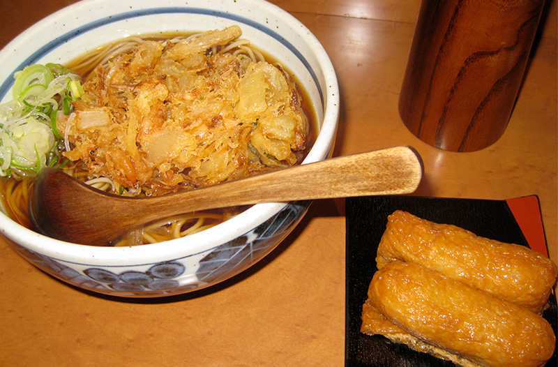 A top-down view of a white ceramic bowl filled with buckwheat soba noodles in a dark soy-based broth, topped with a large, golden-brown vegetable tempura kakiage and fresh green onions, served alongside two pieces of inarizush