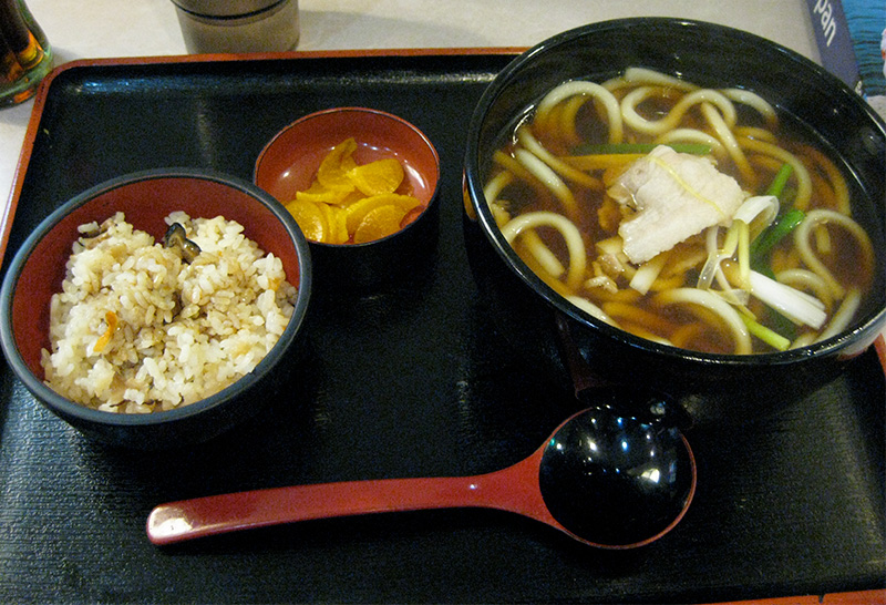 A Japanese udon set meal served on a black tray, featuring a large bowl of thick wheat noodles in a clear broth topped with sliced fish cake and green onions, accompanied by a bowl of mixed seasoned rice (takikomi gohan), a small dish of yellow pickled radish (takuan), and a traditional red and black soup spoon.