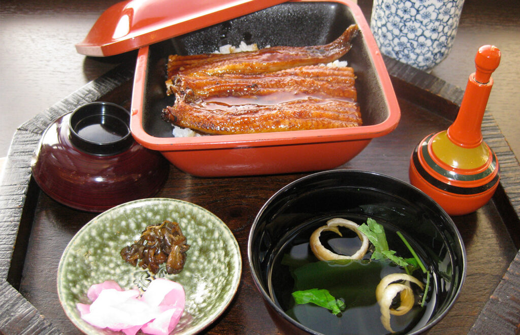 A traditional Japanese unagi-no-kabayaki meal featuring grilled eel glazed in a dark savory sauce served over a bed of white rice in a rectangular red and black lacquer box (unaju), accompanied by a side of green pickles and a bowl of clear dashi soup.