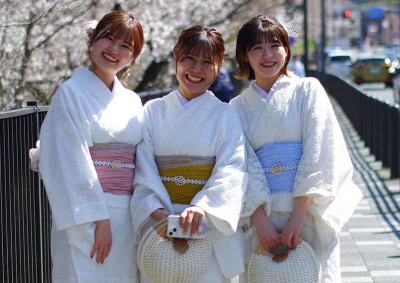 Three women dressed in colorful traditional kimonos and yukata standing together on a stone bridge in Kyoto, smiling and posing for a photo with historic wooden buildings in the background.