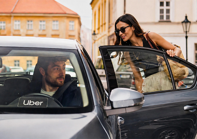 A woman in sunglasses and a denim jacket leans into the open door of an Uber, smiling and talking to the driver, while another woman is already seated inside. The car is on a city street lined with European-style architecture.