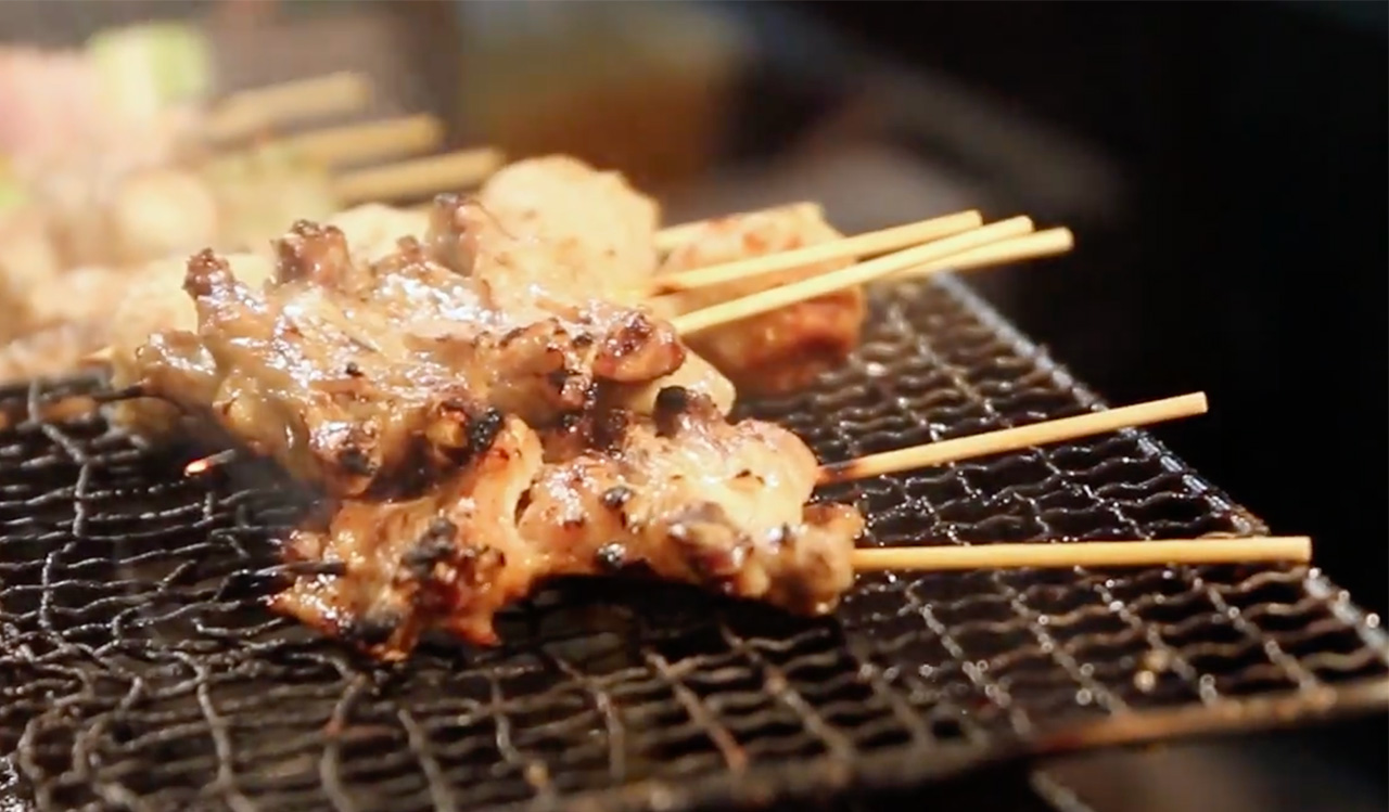 Close up of several chicken yakitori skewers cooking over a traditional charcoal grill with glowing embers and light smoke