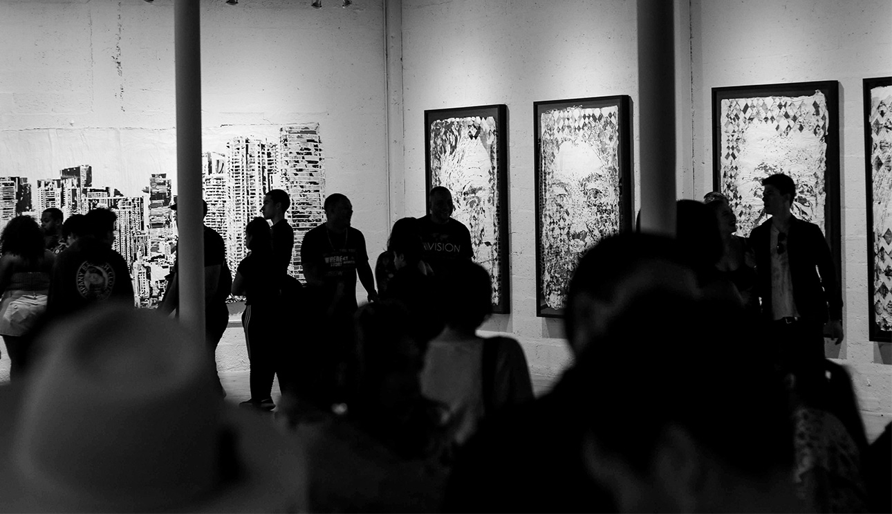 A black and white photograph showing the interior of an art gallery during Art Basel, with several people viewing three large, vertically-oriented framed artworks on a white wall.