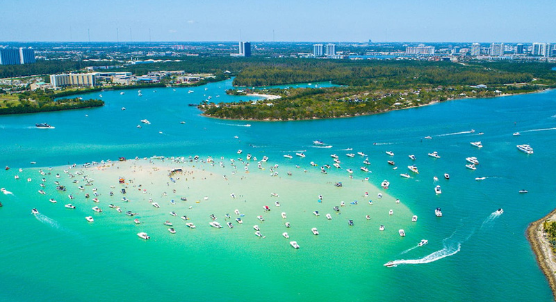 An aerial view of Biscayne Bay with several white boats anchored in the clear, turquoise water near a lush green shoreline under a bright blue sky.