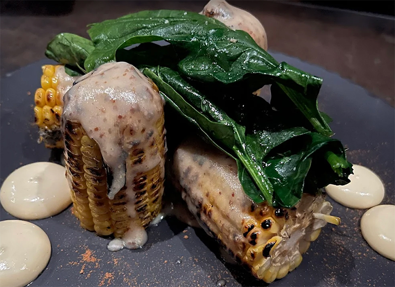 A close-up shot of a gourmet Mexican street corn (elote) from CarbónCabrón, featuring a grilled corn cob heavily coated in creamy sauce and crusted with cheese, topped with a large, vibrant green leaf and served on a dark, textured plate.