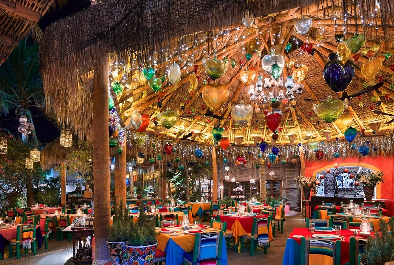 A vibrant, upscale restaurant interior at night featuring traditional Mexican decor, glowing hanging lanterns, and colorful table settings under a large palapa-style thatched roof at Edith's in Cabo San Lucas.