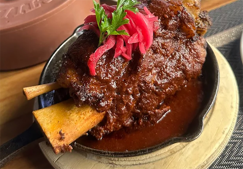 A close-up of a tender, slow-cooked beef rib glazed in a rich savory sauce, served on a wooden board at El Peregrino in Cabo San Lucas.