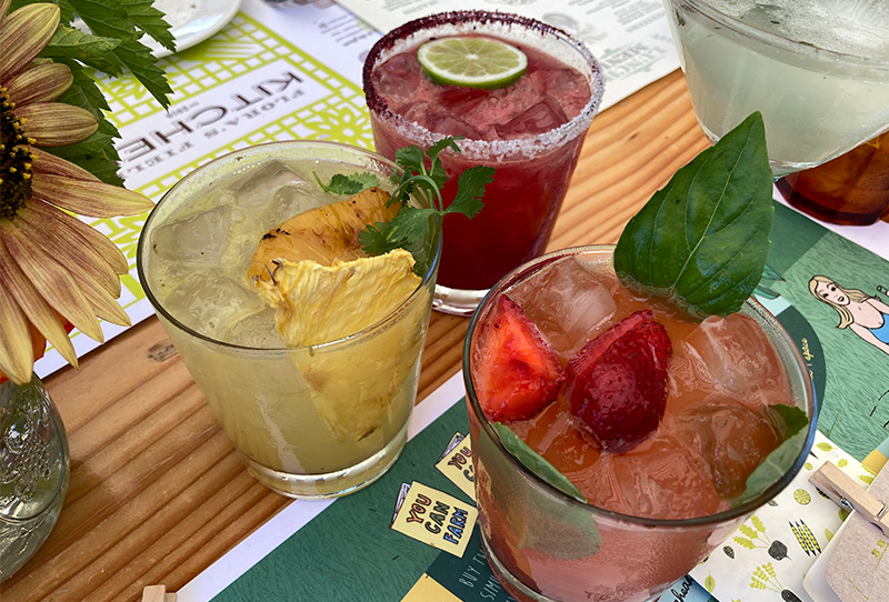 Two colorful farm-to-table cocktails, one yellow and one red, garnished with fresh herbs and fruit, sitting on a wooden table at Flora Farms in Los Cabos.