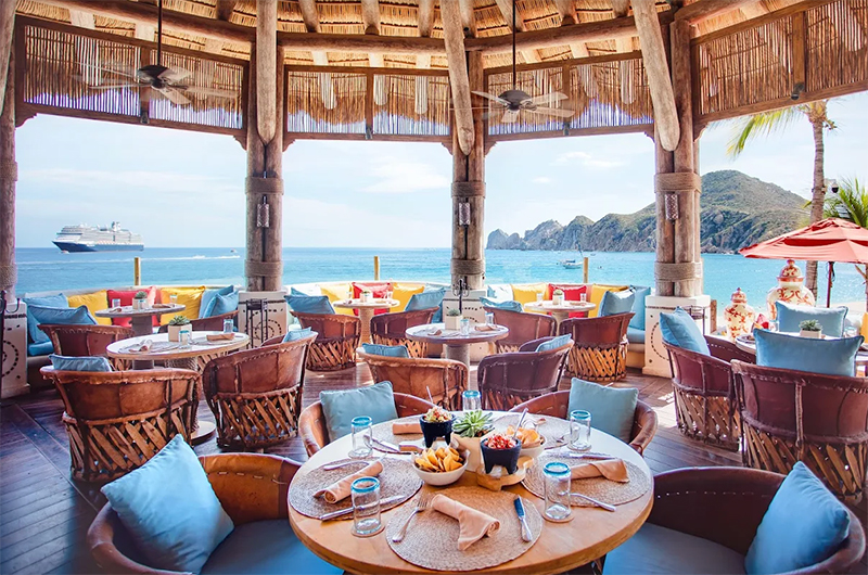 A panoramic view of a luxurious beachfront restaurant terrace at night, featuring white-clothed tables, elegant lighting, and palm trees overlooking the calm ocean and the distant silhouette of the Land's End rock formations.