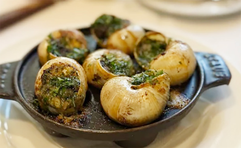 A close-up of a classic French escargot dish at LPM Restaurant & Bar in Miami, featuring snails baked in their shells with a vibrant green garlic and herb butter, served in a traditional dark ceramic escargot dish