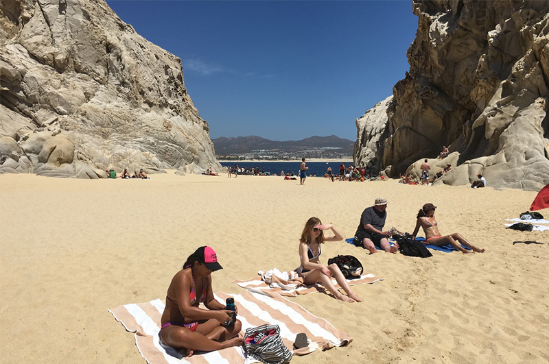 A view of the secluded Lover’s Beach in Cabo San Lucas, nestled between towering granite rock formations with golden sand and clear turquoise water under a bright blue sky.