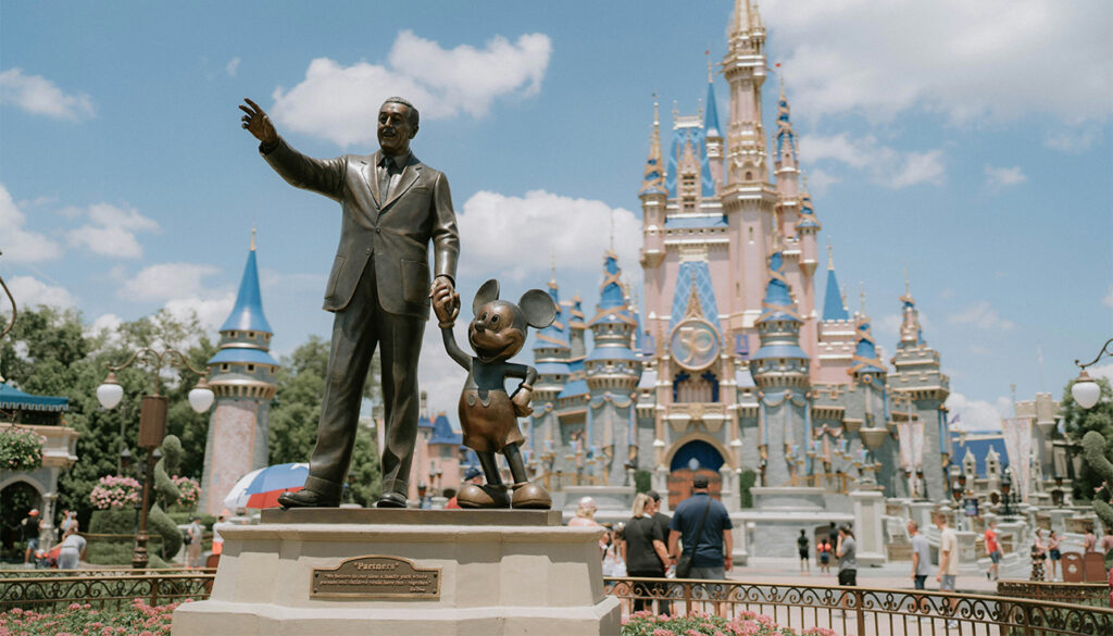 A wide view of the Magic Kingdom at Walt Disney World, featuring the iconic "Partners" statue of Walt Disney and Mickey Mouse in the foreground, with the spires of Cinderella Castle rising behind them under a soft sky.