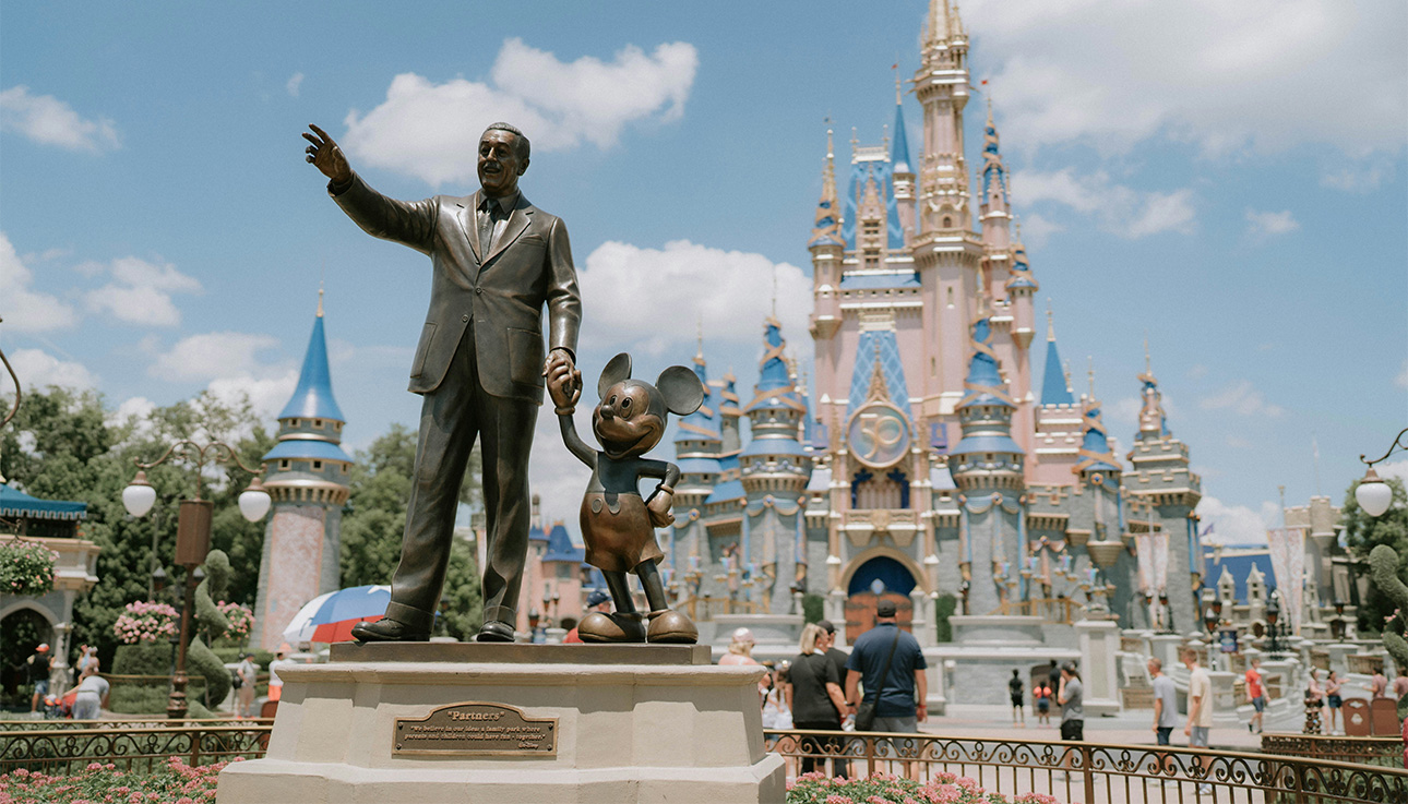 A wide view of the Magic Kingdom at Walt Disney World, featuring the iconic "Partners" statue of Walt Disney and Mickey Mouse in the foreground, with the spires of Cinderella Castle rising behind them under a soft sky.