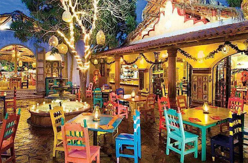 A colorful and festive open-air restaurant exterior at night, featuring traditional Mexican architecture, bright blue and yellow wooden chairs, and decorative hanging lanterns illuminating a lush courtyard at Mi Casa in Cabo San Lucas.