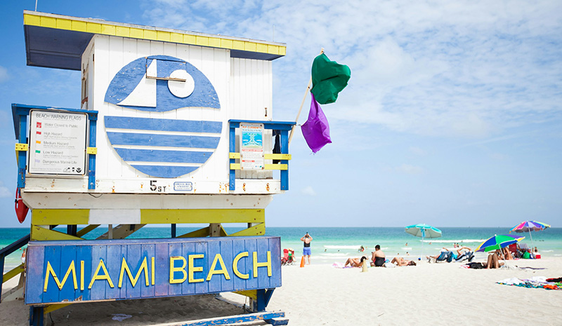 A iconic Miami Beach lifeguard tower painted with light blue and yellow accents, featuring a circular window and "MIAMI BEACH" lettering, situated on a bright sandy shore with a purple flag flying against a clear sky.