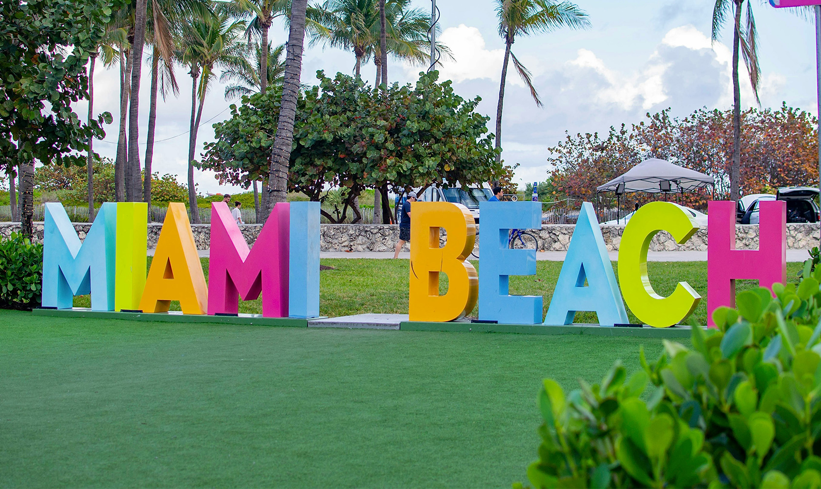 A colorful "Miami Beach" sign with large, block letters in vibrant colors like yellow, pink, and green, set against a backdrop of lush palm trees and a bright blue sky