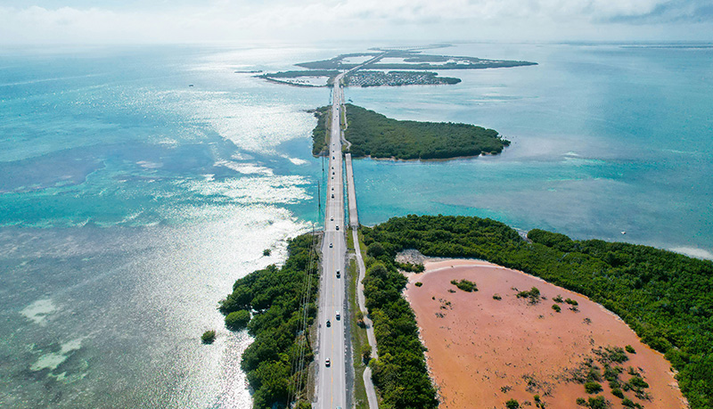 An aerial perspective of the Seven Mile Bridge in the Florida Keys, showing the long, modern highway stretching across a vast expanse of light blue and turquoise tropical water under a soft, hazy sky.