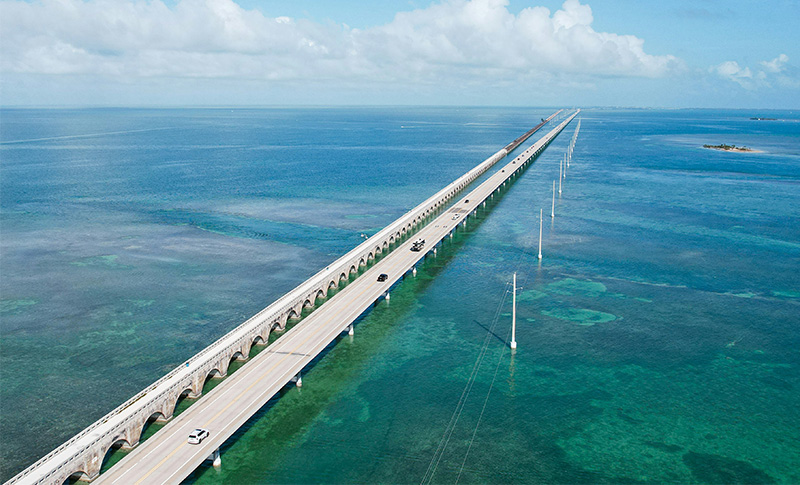An aerial view of the Seven Mile Bridge in the Florida Keys, showing the long, modern highway stretching across a vast expanse of light blue and turquoise tropical water under a soft, hazy sky.