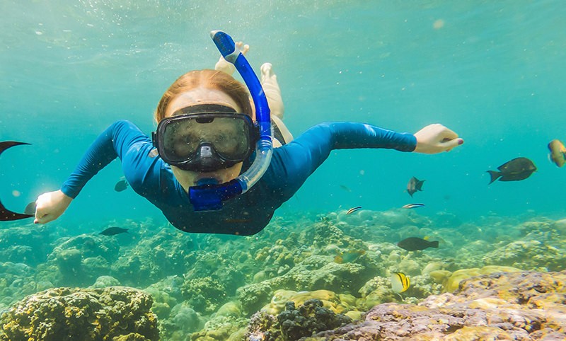 A person wearing a blue rash guard, mask, and snorkel swims through clear turquoise water above a vibrant coral reef teeming with small tropical fish.