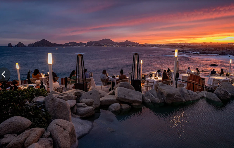 A dramatic cliffside restaurant view at sunset, featuring tiered outdoor dining terraces with white-clothed tables overlooking the calm ocean and the iconic Land's End rock formations under a vibrant orange and purple sky.