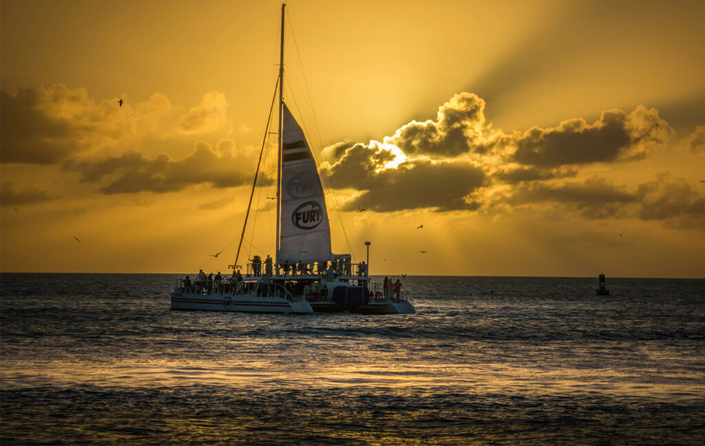 A peaceful sunset view from the water in Key West, featuring a silhouette of a sailing catamaran against a vibrant orange and yellow sky with soft clouds reflecting on the calm ocean.