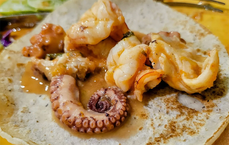 A close-up of a fresh seafood taco from Tacos Gardenias, featuring breaded shrimp or fish topped with shredded cabbage, fresh salsa, and a squeeze of lime on a traditional corn tortilla