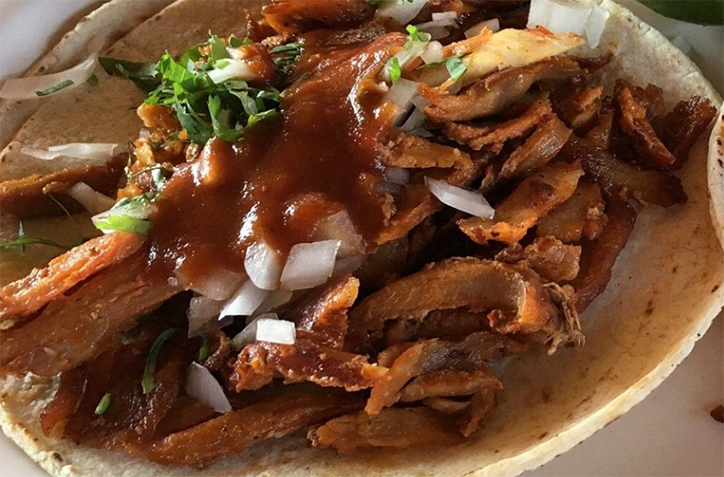 A close-up of a traditional Mexican taco al pastor from Taqueria El Fogon, featuring thinly sliced marinated pork topped with fresh cilantro, onions, and a slice of pineapple on a corn tortilla.