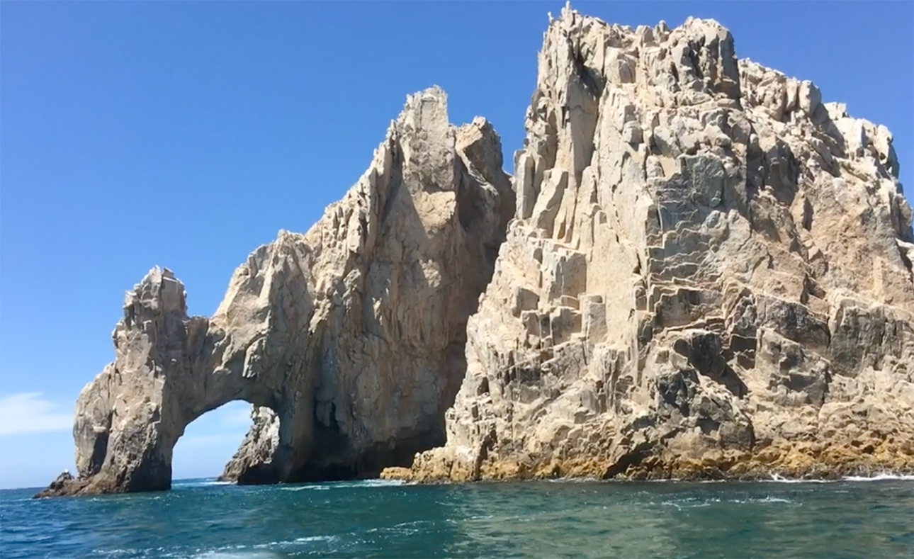 A view of the rugged, golden limestone Arch of Cabo San Lucas (El Arco) rising from the turquoise waters of the Sea of Cortez under a clear blue sky.